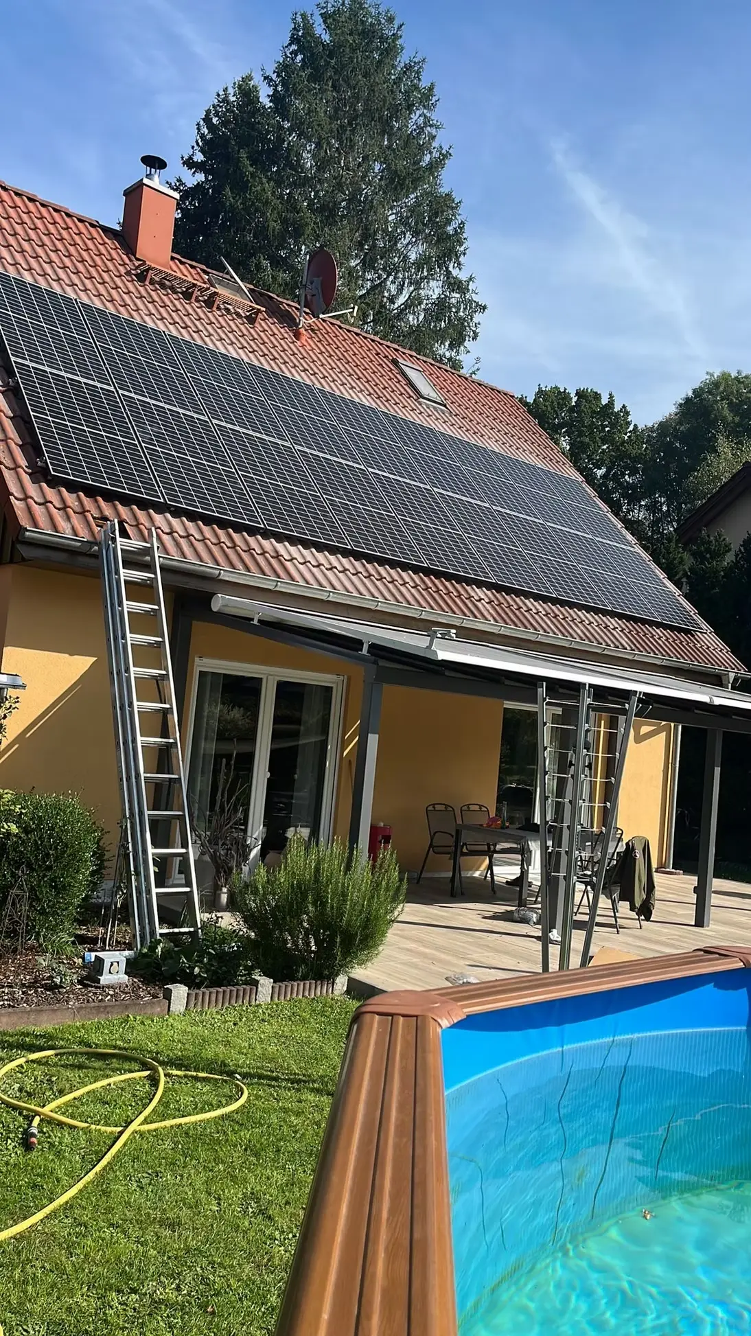Solarpaneele auf dem Dach eines Hauses mit einem Garten und einem Pool im Vordergrund. Eine Leiter steht an der Hauswand, während eine Terrasse mit Möbeln im Hintergrund zu sehen ist. Klarer Himmel und Bäume umgeben die Szene.