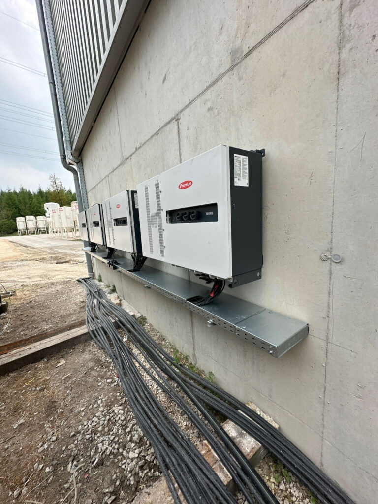 Bild einer Wand mit mehreren Fronius-Wechselrichtern, die auf einer Metallablage installiert sind. Kabel verlaufen entlang des Bodens. Die Umgebung zeigt eine Baustelle mit unbefestigtem Boden und Bäumen im Hintergrund.